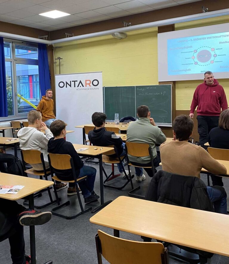 Vortrag von ONTARO in einem Klassenzimmer vor Schülern mit Präsentationsfolie und Roll-up-Banner.
