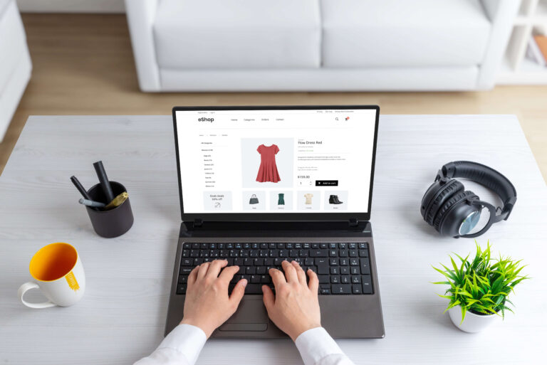 Ein geöffneter Laptop auf einem Schreibtisch, zwei Hände schreiben auf der Tastatur auf dem Bildschirm ist ein Fashion Online Shop zu sehen.