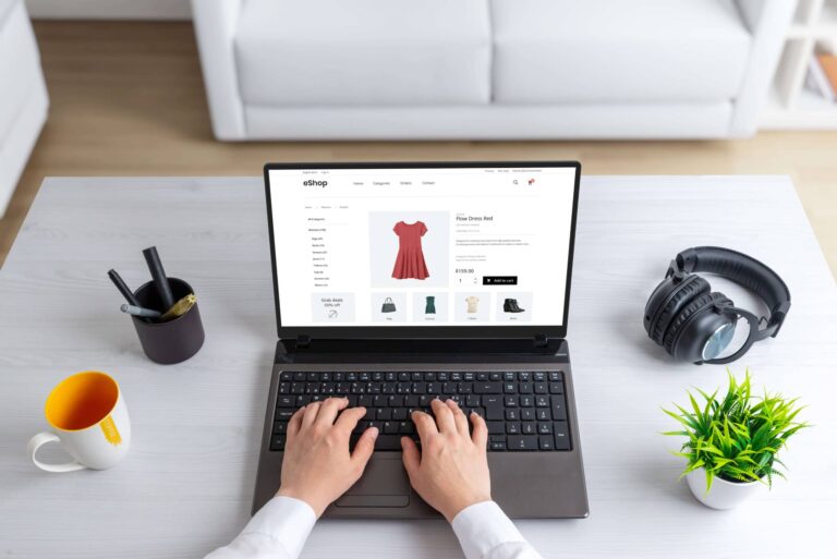 Ein geöffneter Laptop auf einem Schreibtisch, zwei Hände schreiben auf der Tastatur auf dem Bildschirm ist ein Fashion Online Shop zu sehen.