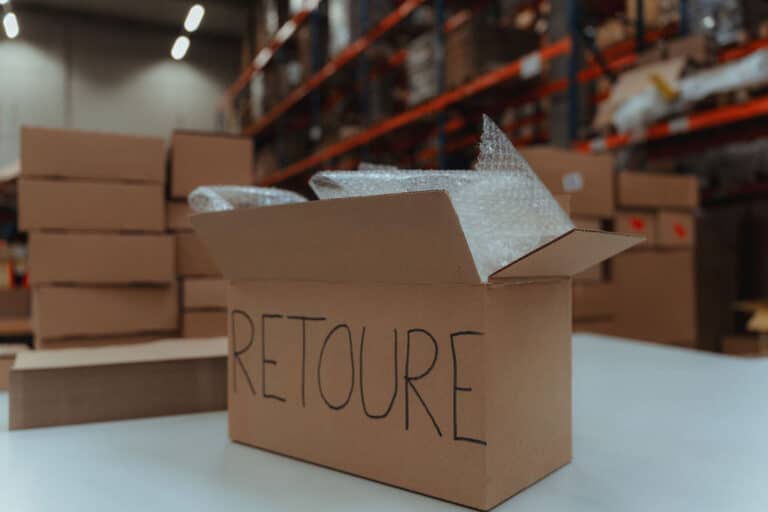 Ein geöffneter Versandkarton mit der Aufschrift 'RETOURE' und Schutzfolie auf einem Arbeitstisch im Logistikzentrum.