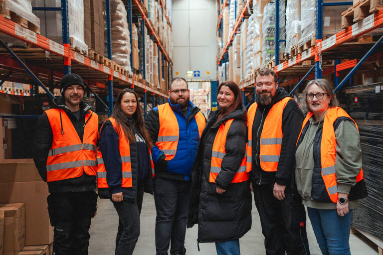 Sechs Lagermitarbeiter:innen mit Sicherheitsweste in einem Lager mit Regalen im Hintergrund