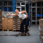 Eine lächelnde Mitarbeiterin im Logistikzentrum balanciert gut gelaunt in einer Surf-Pose auf einem Rollbrett. Im Hintergrund stapeln sich Kartons auf Paletten und Lagerregale.